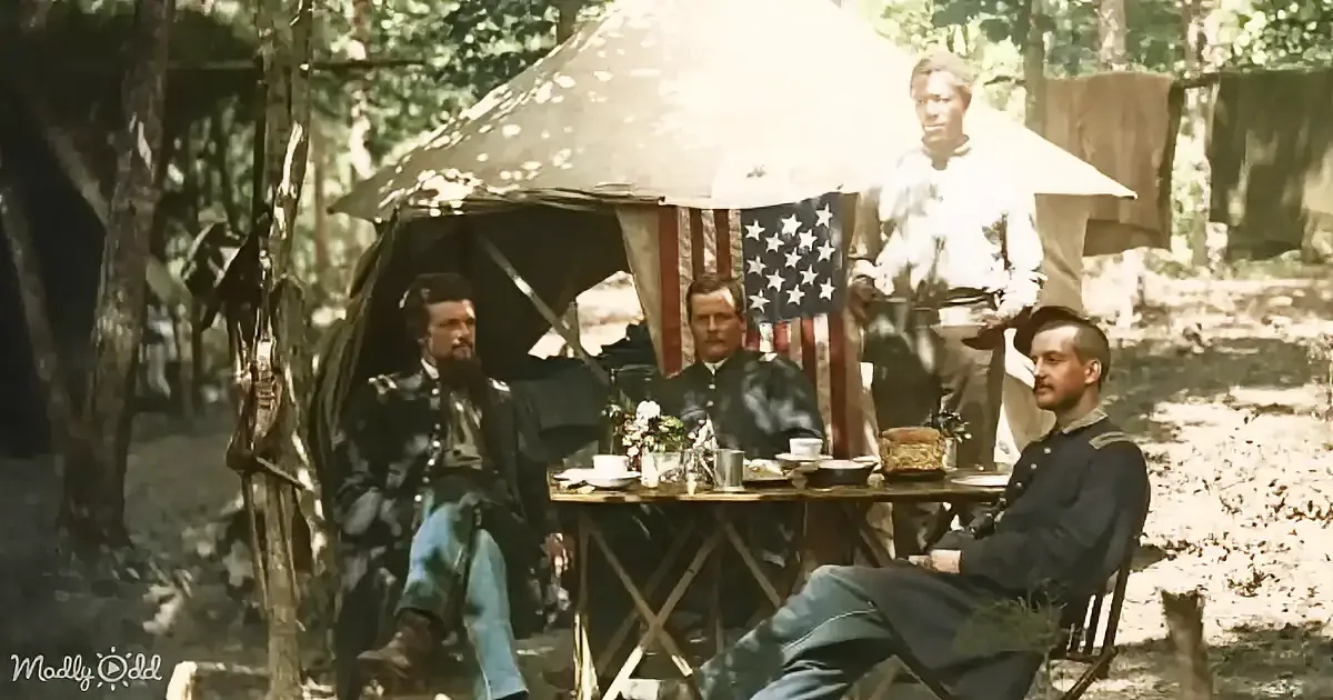 Three men in military uniforms sit at a table with an American flag in a forested campsite.