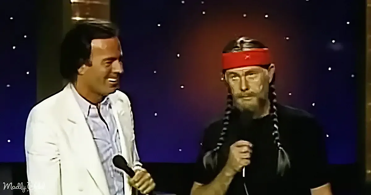 A man in a white suit holds a microphone, smiling, next to another man in a black outfit with braids and a red headband.