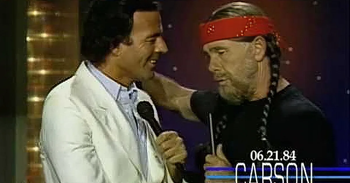 Two men, one in white jacket, the other in black with red headband, stand together, one with hand on shoulder.