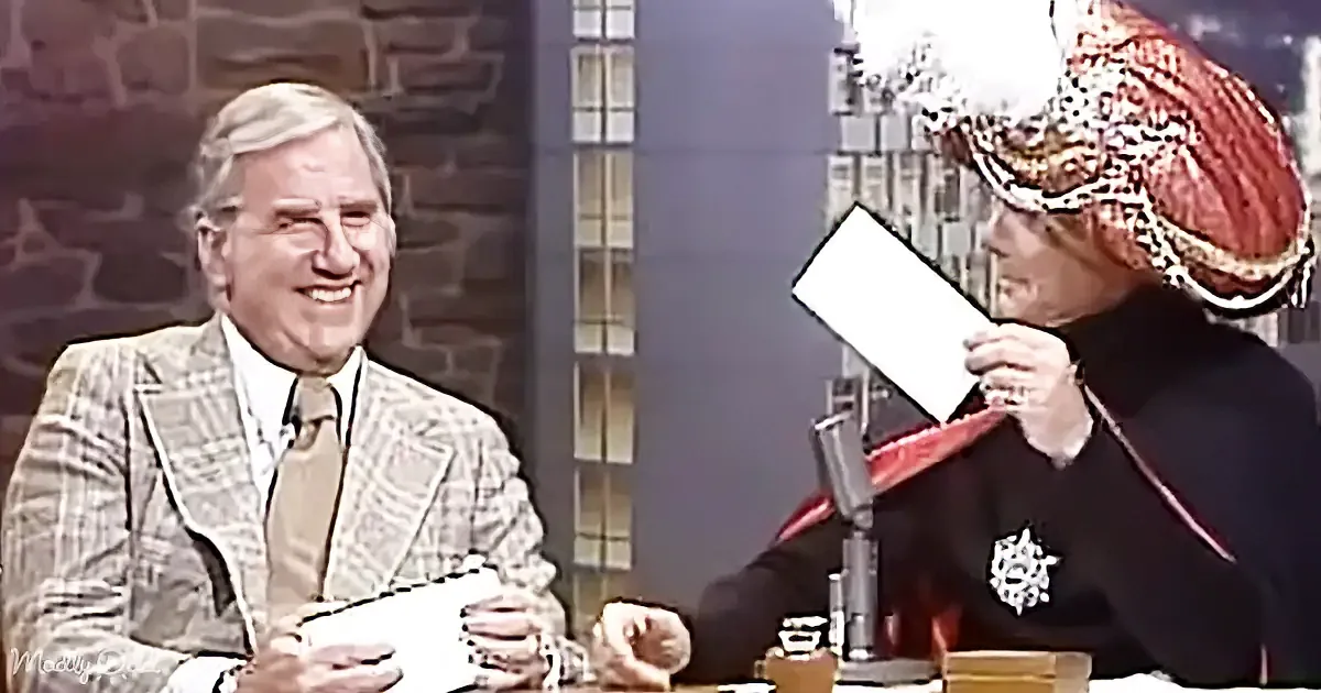 A man in a plaid suit smiles while a woman in a red hat reads from a paper.