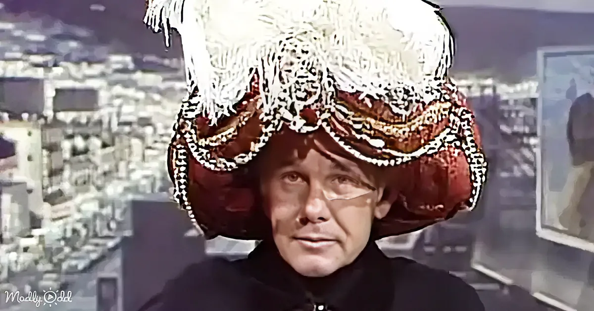A person wearing an elaborate, colorful headdress with fringe and beads stands in front of a cityscape.