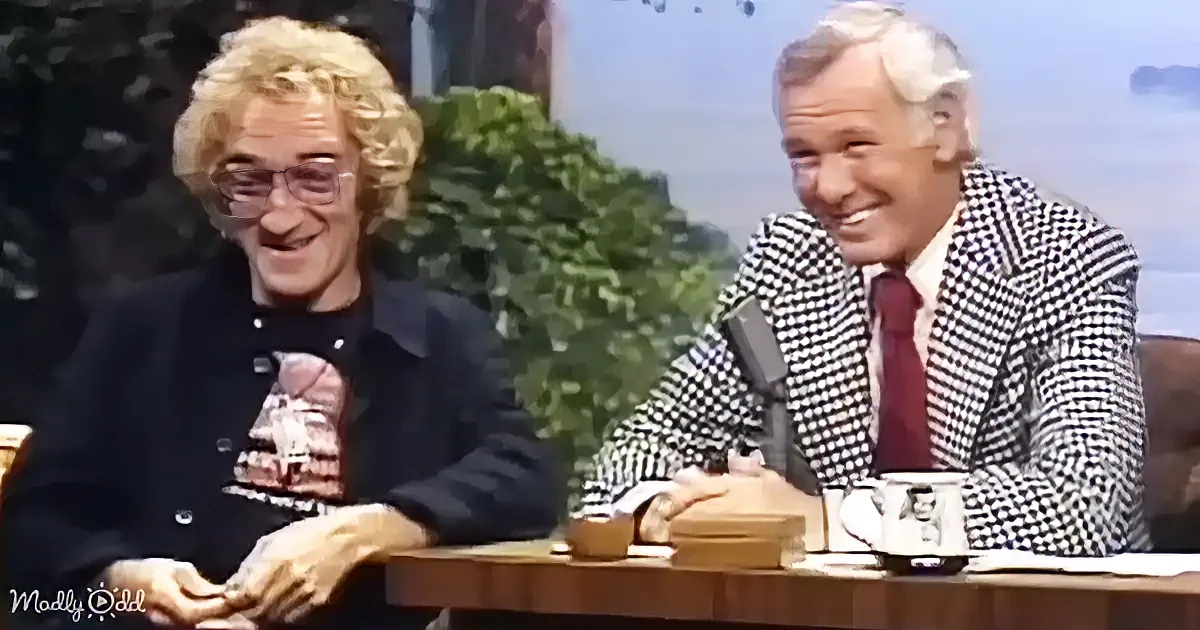 Two men, one in a black jacket and the other in a checkered suit, are seated at a table with a mug and a small object...