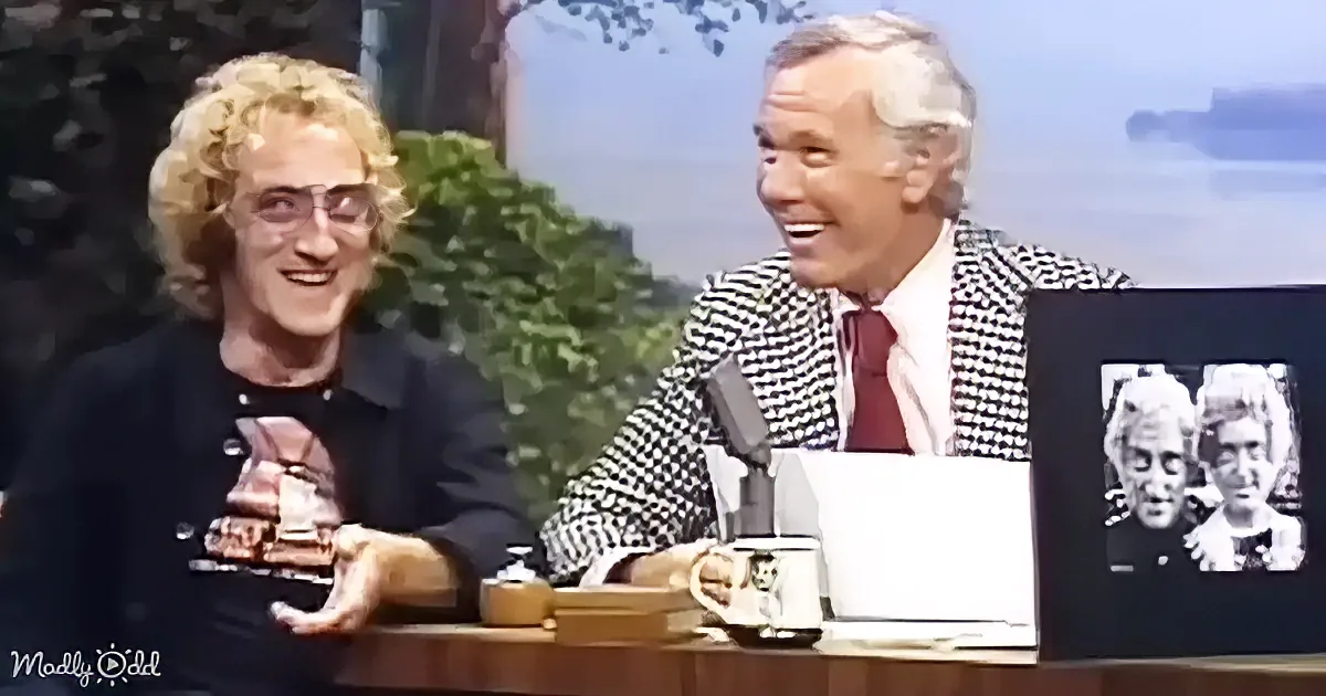 Two men are seated at a table, one in a checkered suit, smiling, with a mug and papers in front of them.