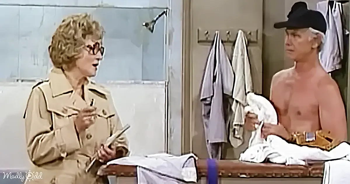 A woman in a beige trench coat holds a book, smiling, while a man in a white robe sits on a bench in a locker room.