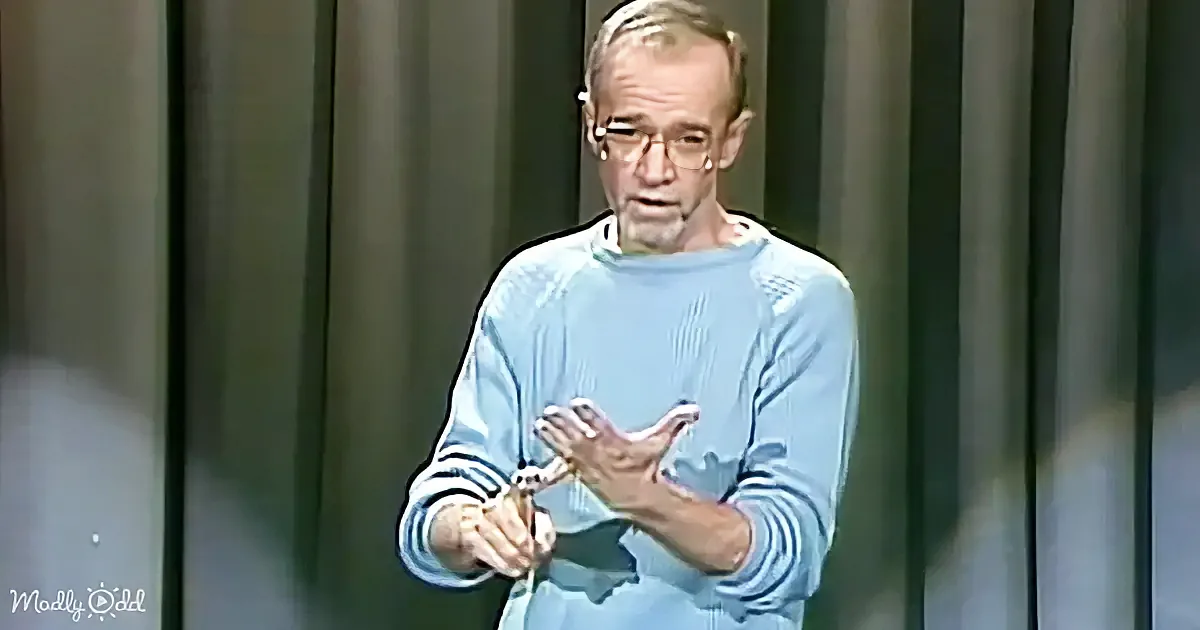 A man in a light blue sweater gestures with his hands while speaking, wearing glasses.