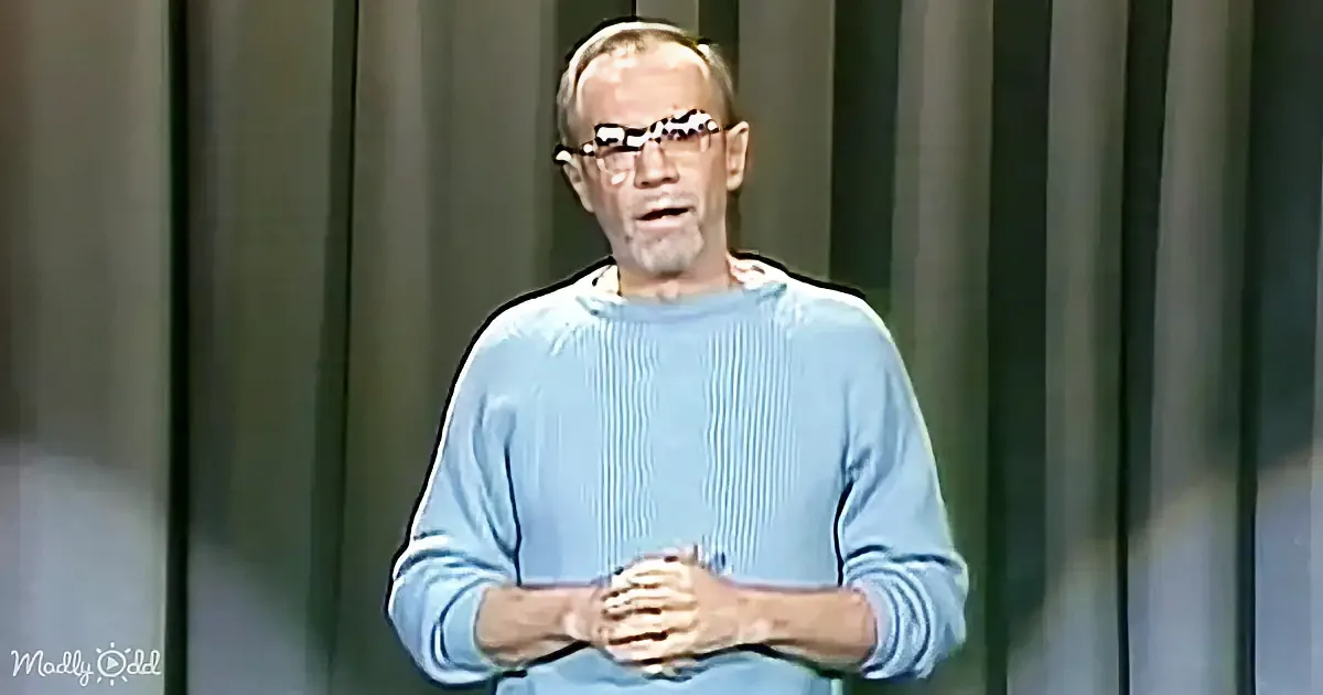 An older man in a light blue sweater stands with hands clasped, wearing glasses, against a dark curtain backdrop.