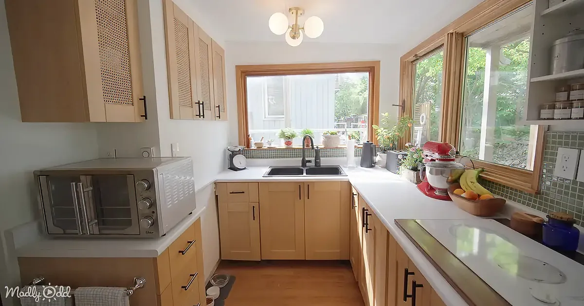 A cozy kitchen with light wood cabinets, a sink, a microwave, and a window with plants and a view of greenery outside.