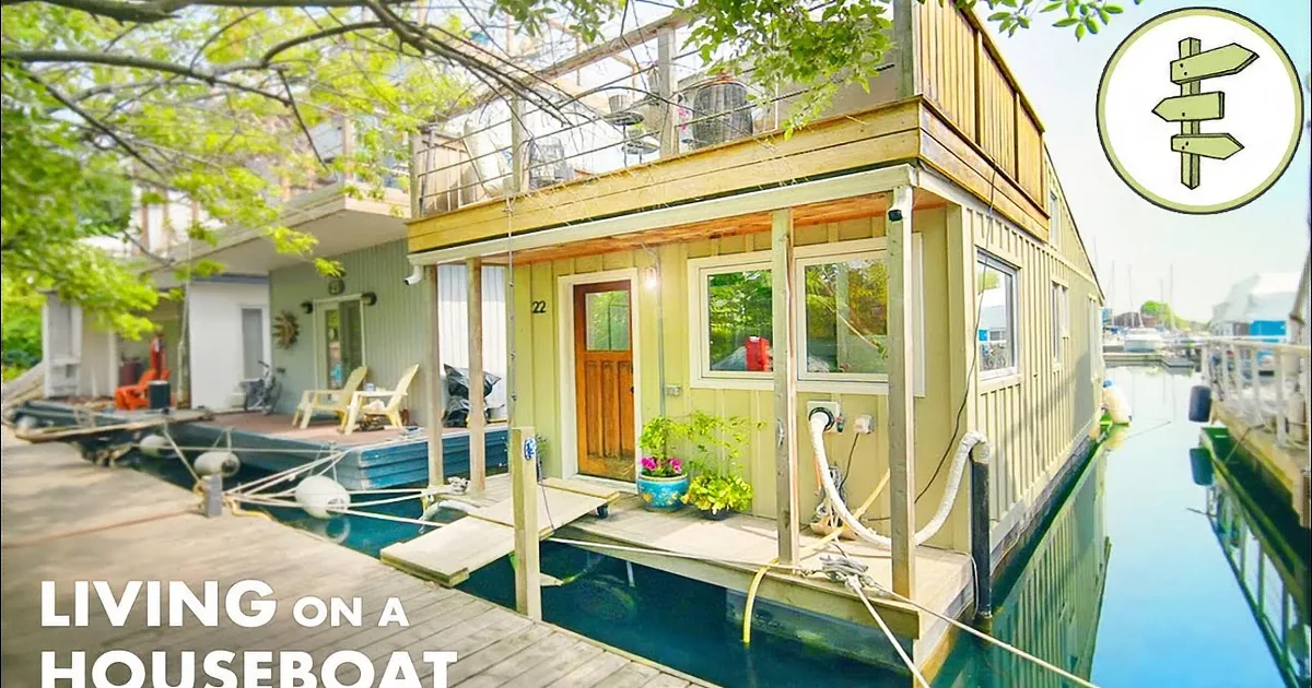 A houseboat with a wooden deck, potted plants, and a small dock is moored in a calm waterway.