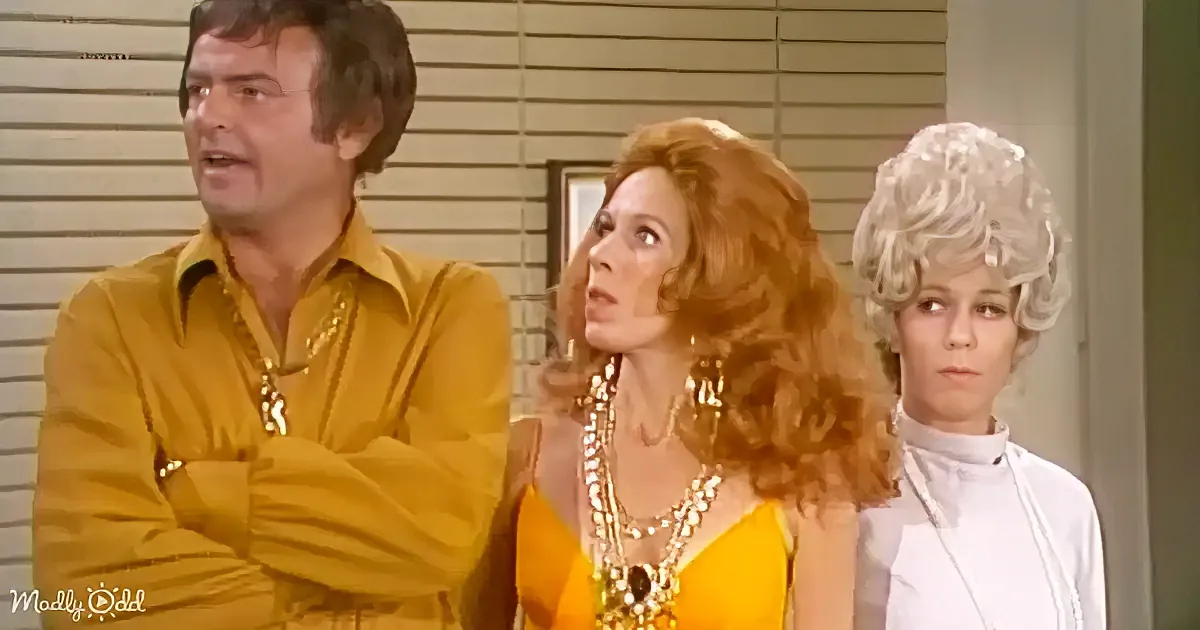 A man in a yellow shirt stands with two women, one in a yellow dress and the other in white, in a room with blinds.
