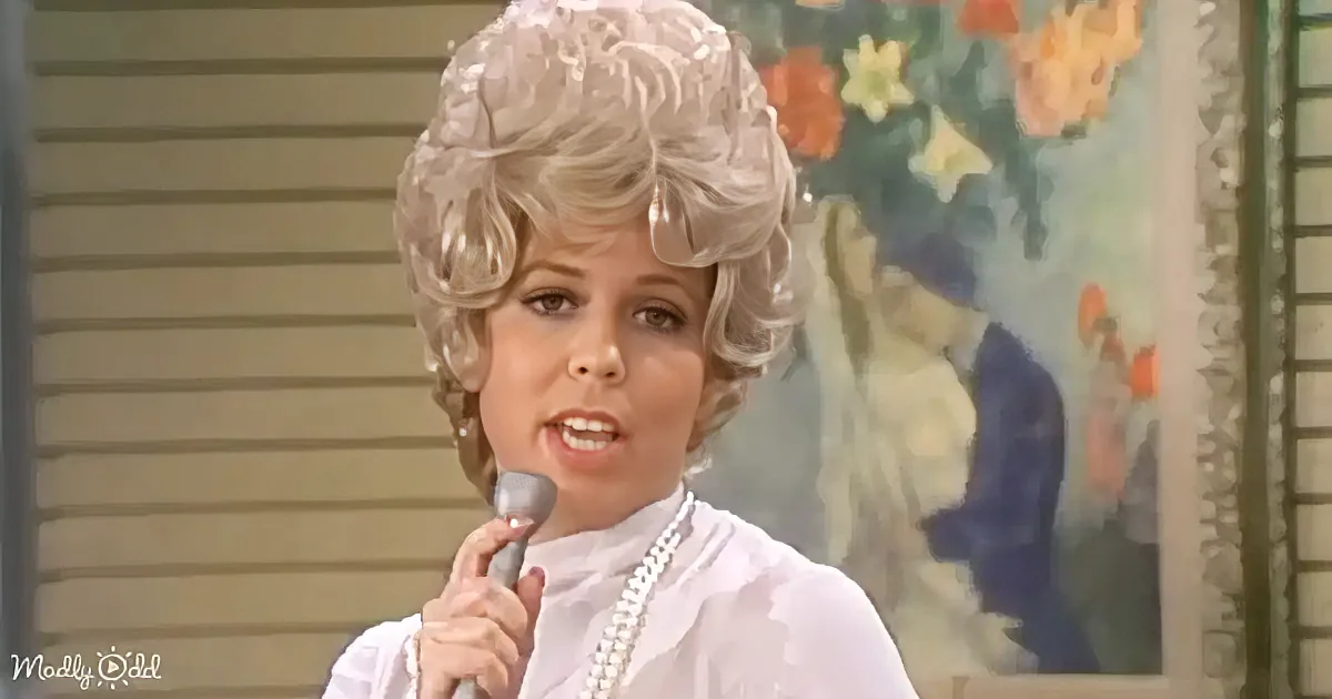 A woman with a voluminous blonde wig and pearls speaks into a microphone, wearing a white blouse.