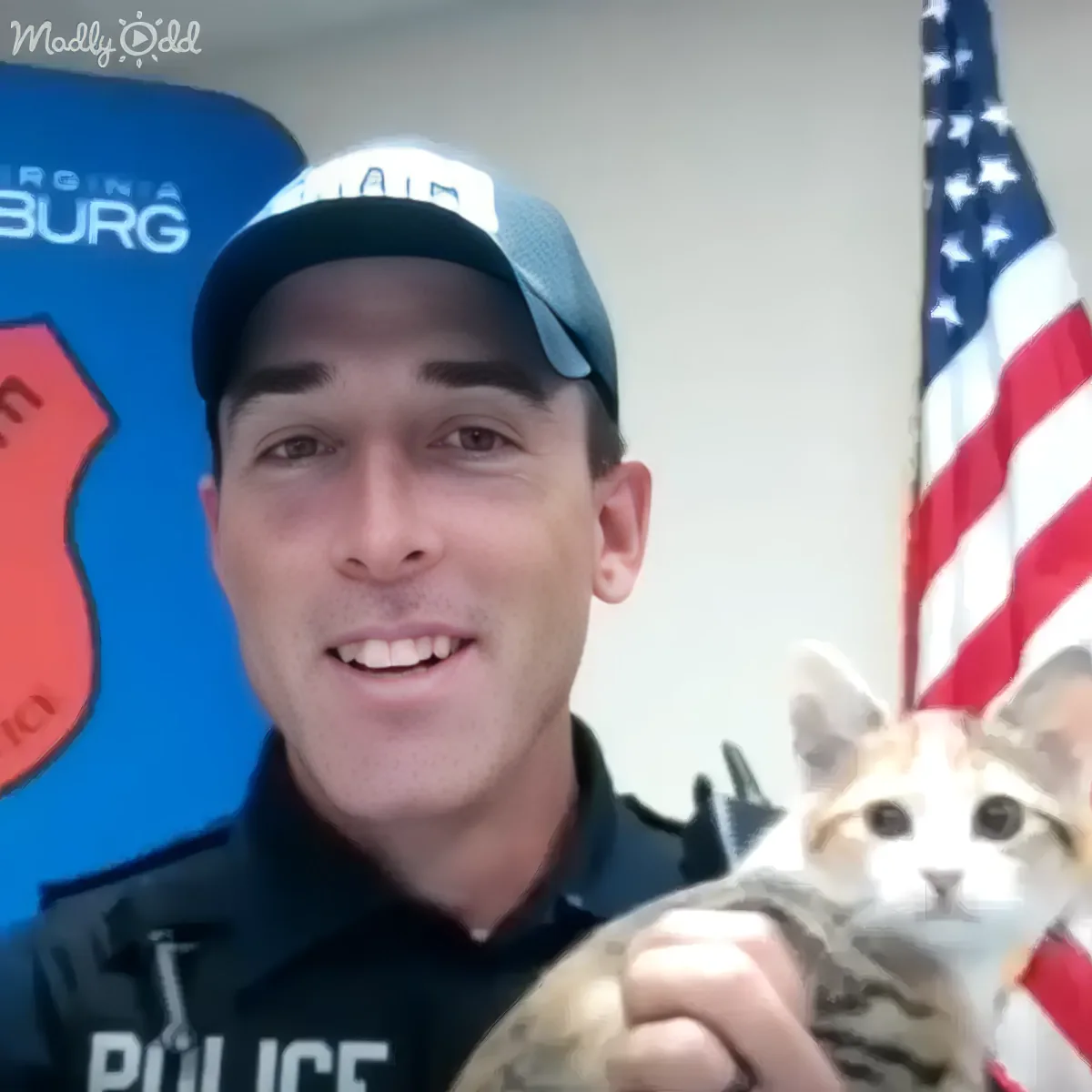 A police officer smiles while holding a cat in front of an American flag and a Virginia sign.