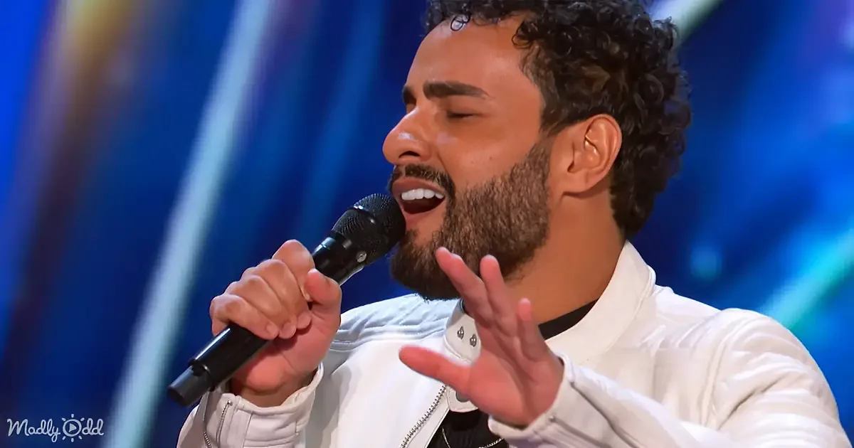 A man with curly hair and a beard sings passionately into a microphone, gesturing with his hand.