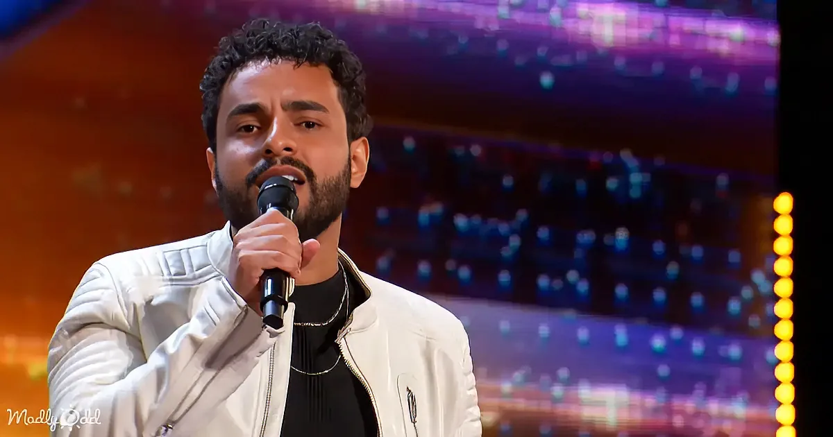 A man in a white jacket sings into a microphone on a stage with colorful lights.