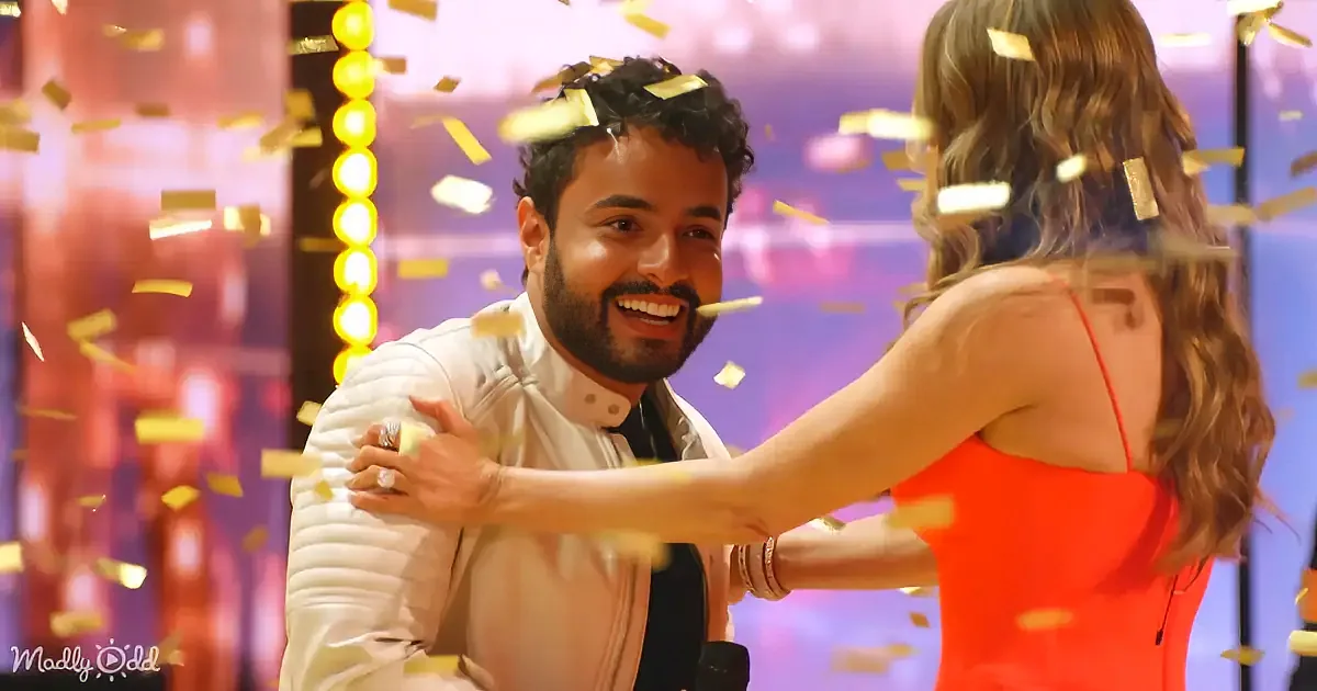 A man in a white jacket smiles as a woman in an orange dress embraces him amidst falling gold confetti.