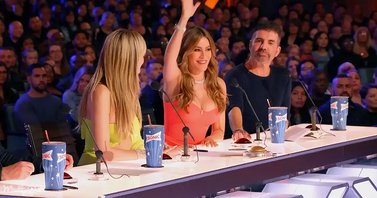 Woman in orange dress raises hand, seated at panel table with microphones and Sonic cups, audience in background.