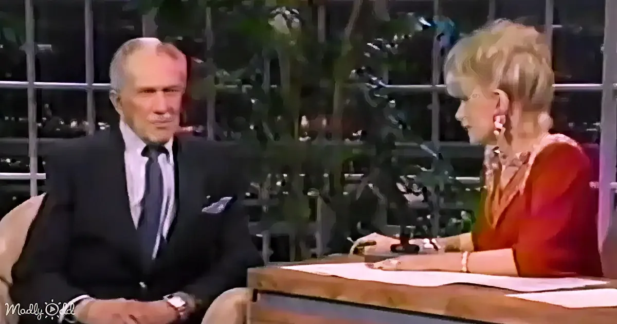 A man in a suit and tie is seated, facing a woman in a red dress, both appearing to be in a talk show setting.