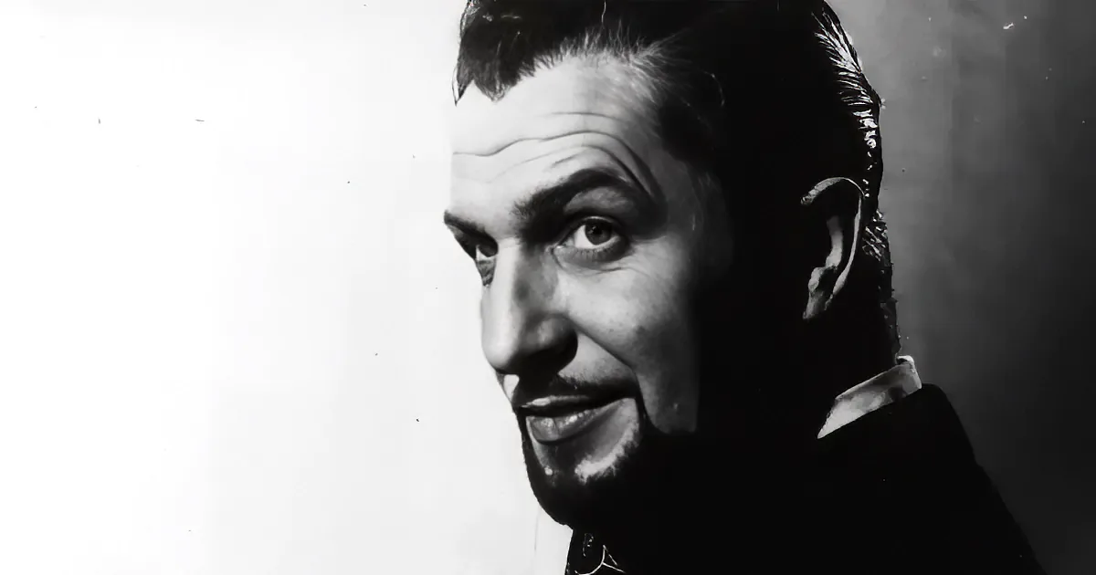 A man with slicked-back hair and a beard, wearing a dark suit, looks to the side with a serious expression.