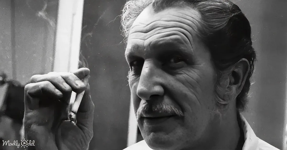 An older man with a mustache holds a cigarette, looking to the side, in a black-and-white photo.