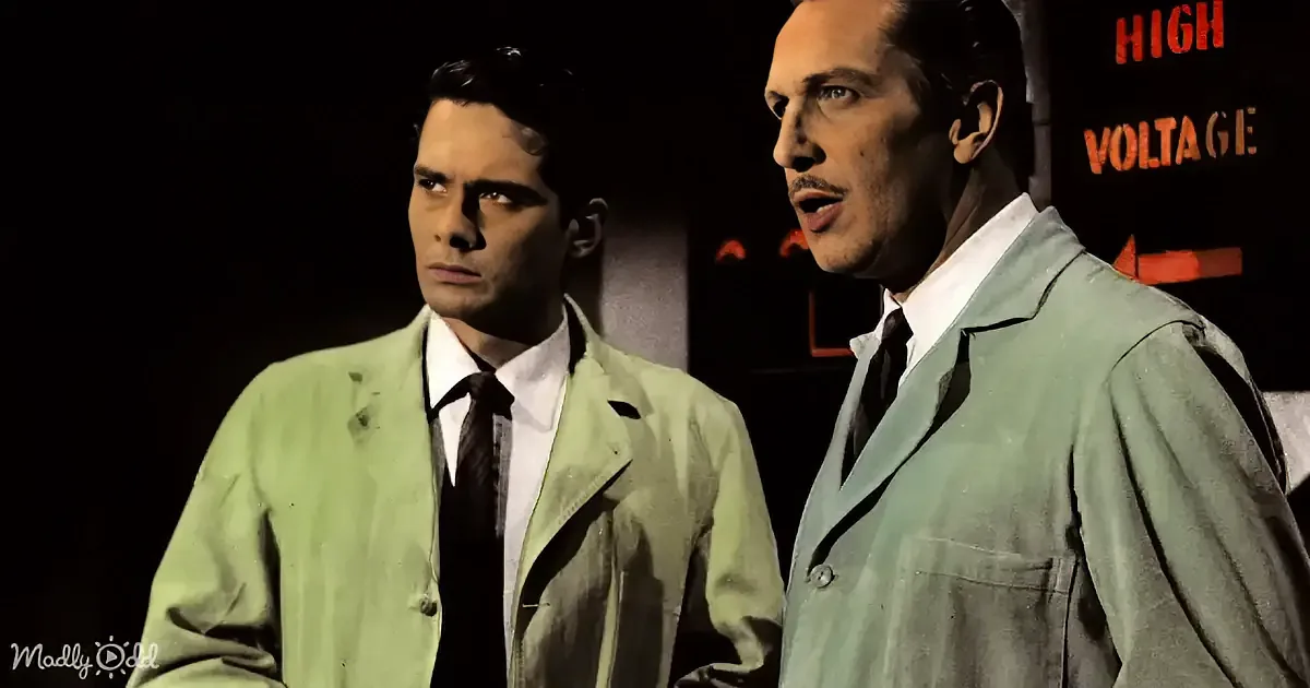 Two men in suits stand in front of a "High Voltage" sign, one speaking, the other listening.