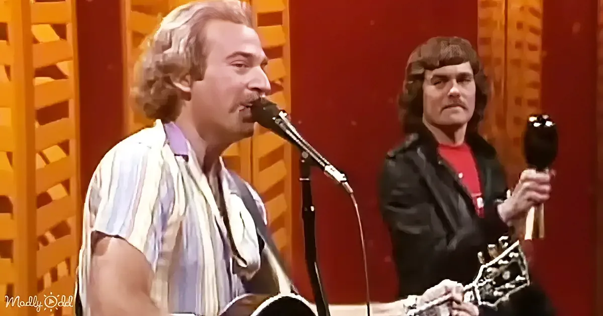A man with curly hair sings into a microphone while another man in a leather jacket holds a tambourine.