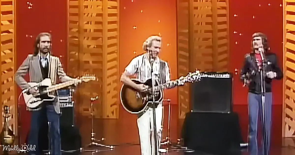 A man in a striped shirt plays guitar on stage with two other musicians.