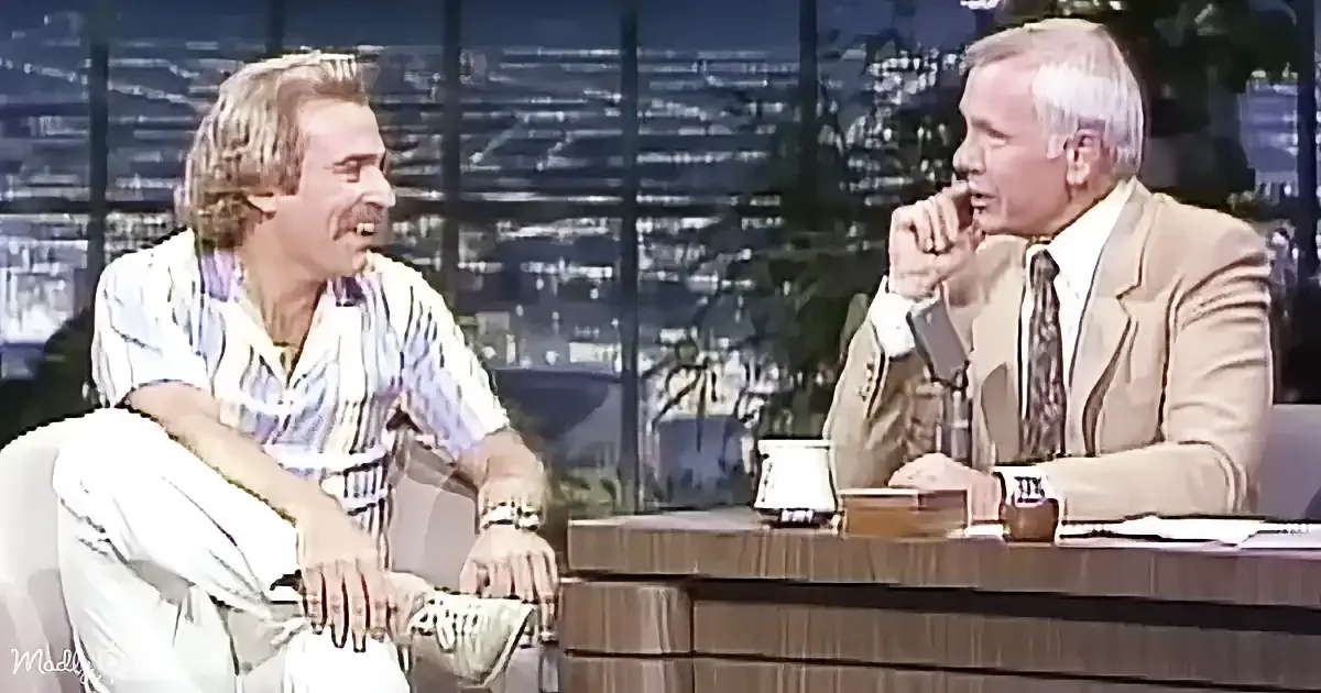 Two men are seated on a talk show set, one in a striped shirt, the other in a suit, engaged in conversation.
