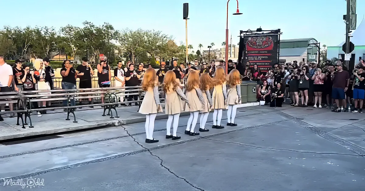 A group of young women in beige dresses and white tights perform in front of a crowd at a public event.