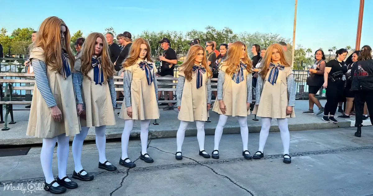 Six identical figures in beige dresses and white tights stand in a row on a concrete surface.