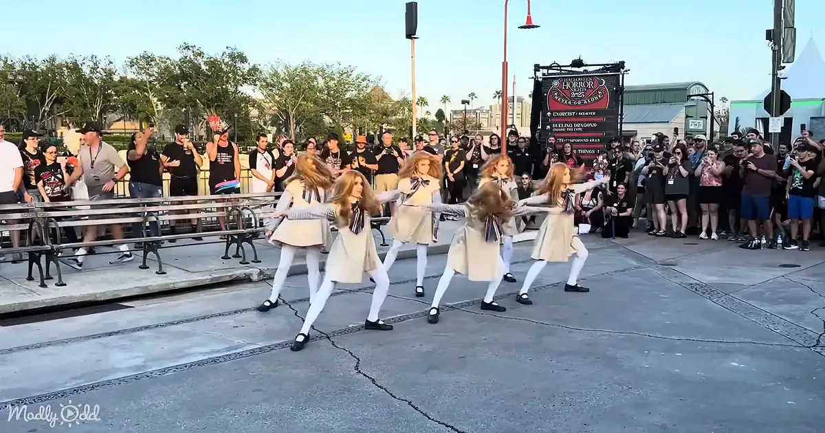 Six performers in beige dresses and white tights dance in front of a crowd at a public event.