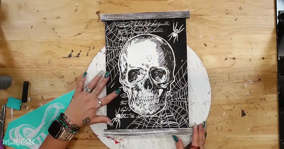 A hand holds a black and white skull illustration with spider webs and text on a wooden table.
