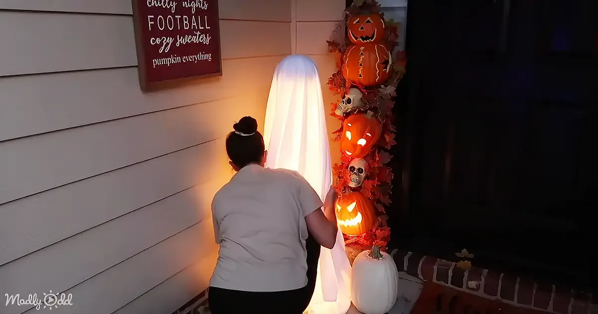 A person adjusts a glowing ghost decoration on a Halloween-themed porch display.