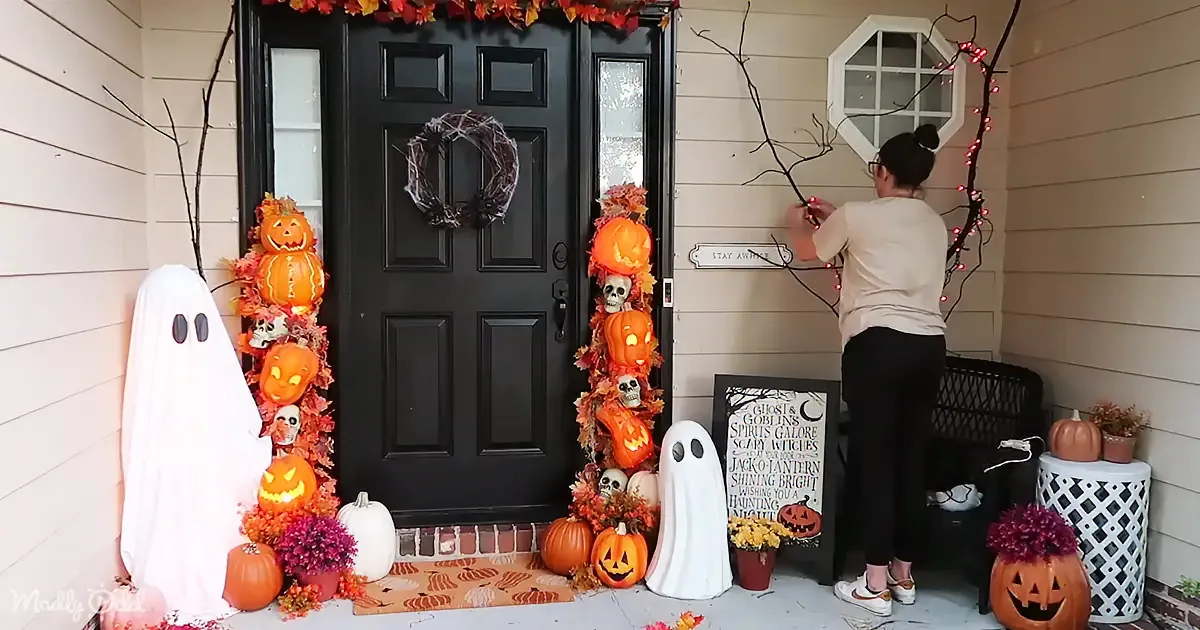 A person decorates a Halloween-themed front porch with pumpkins, ghosts, and lights.