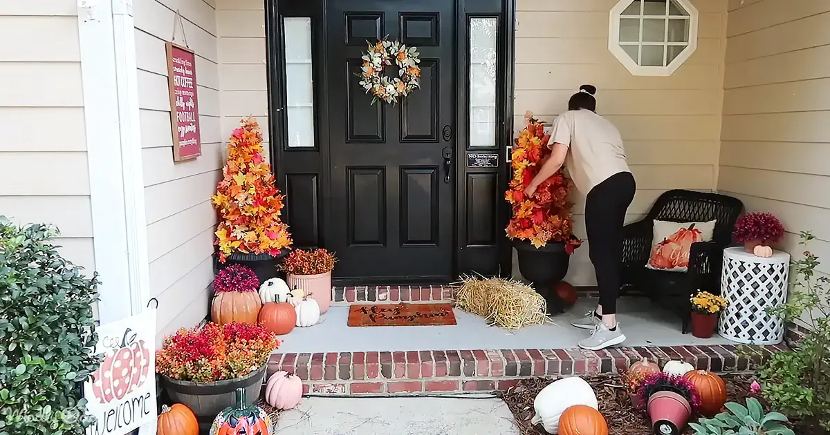 A person decorates a porch with fall-themed items, including wreaths, pumpkins, and flowers.