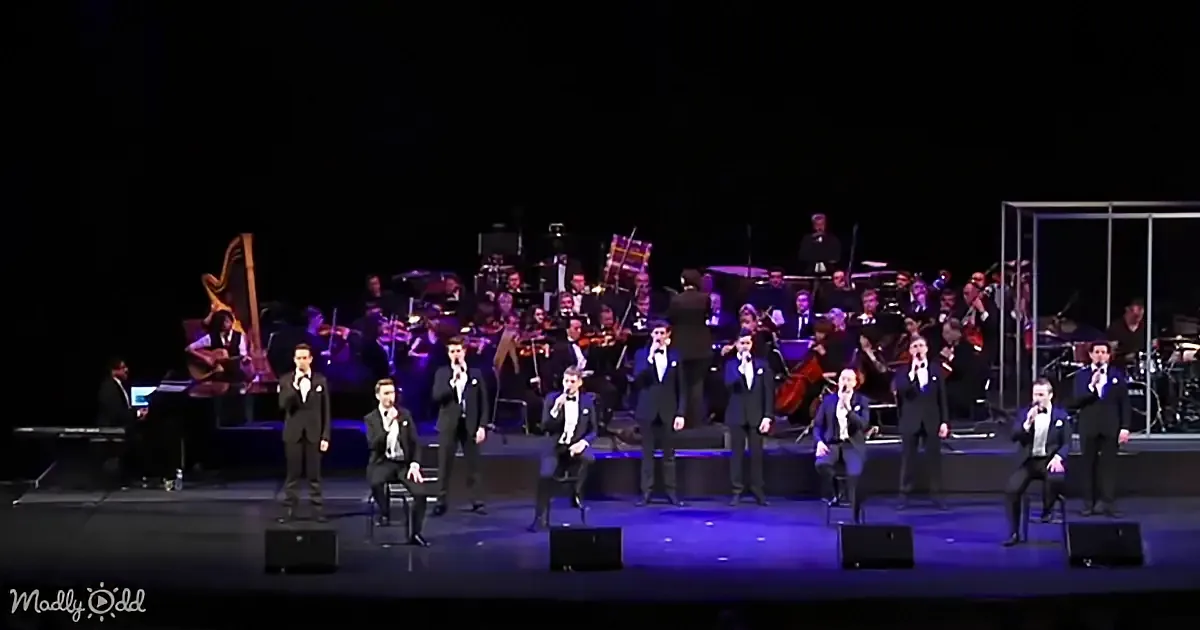 A group of men in suits sing on stage with a large orchestra behind them.