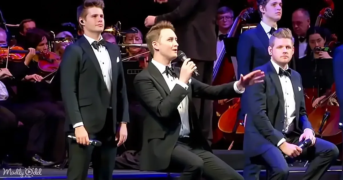 A man in a tuxedo sings into a microphone on stage, surrounded by other men in tuxedos and an orchestra.