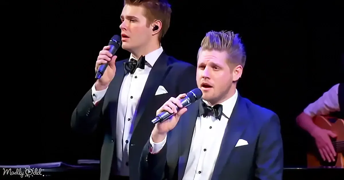 Two men in tuxedos sing into microphones on stage, one playing guitar in the background.