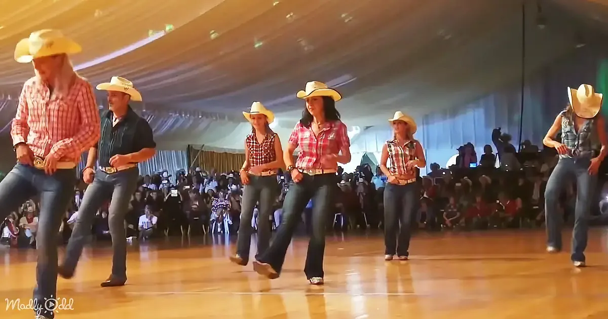Cowboys and cowgirls in plaid shirts and cowboy hats dance on a wooden floor in front of a large audience.