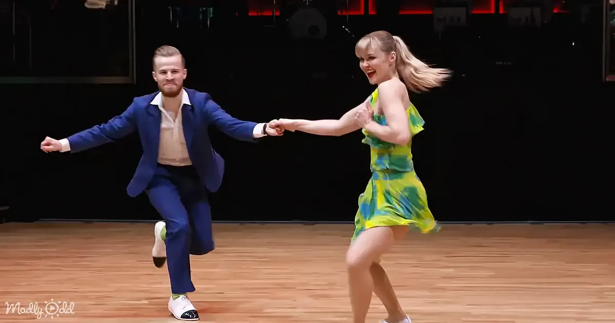 A man and woman are dancing energetically on a wooden floor, holding hands and smiling.