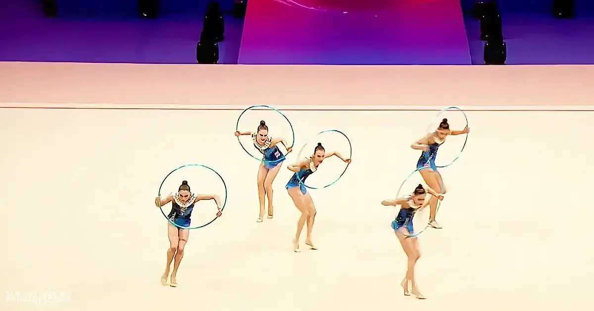 Five gymnasts in blue leotards perform with hoops on a white floor.