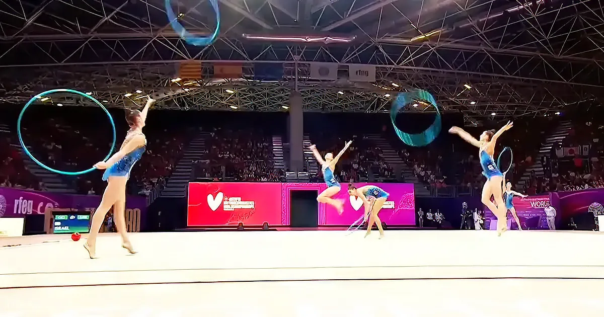Four gymnasts perform with hoops in a synchronized routine on a stage in front of an audience.