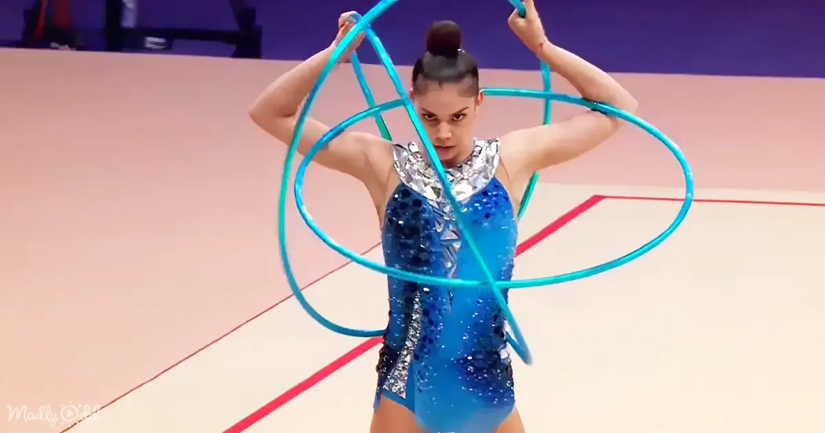 A gymnast in a blue sequined leotard holds two turquoise hula hoops above her head on a gym floor.