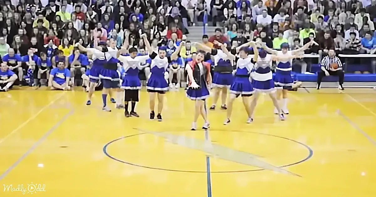 Cheerleaders in blue and white uniforms perform on a basketball court with a crowd watching.