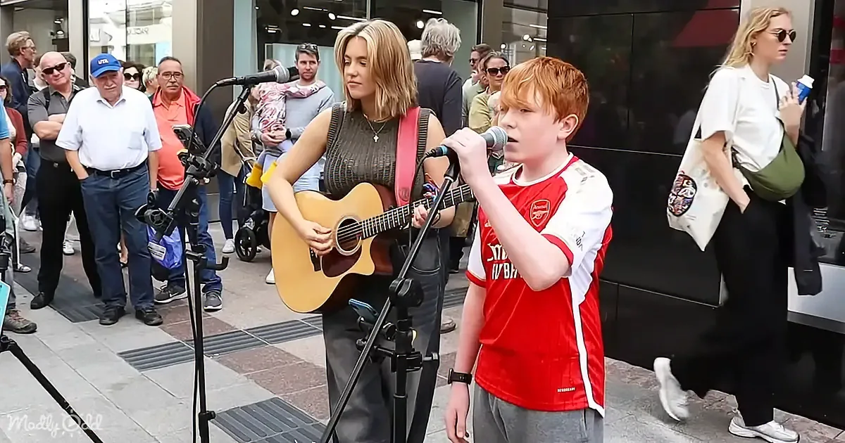 A woman plays guitar while a boy sings into a microphone, surrounded by an audience on a busy street.