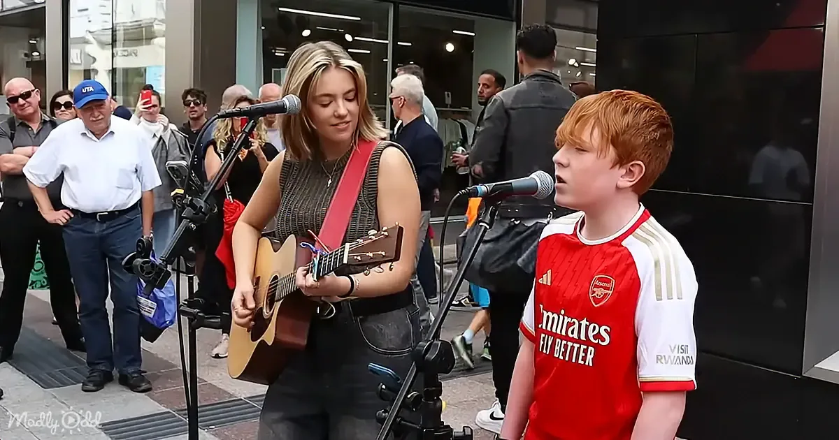 A woman plays guitar while a boy sings in front of a crowd, both on a street performance stage.