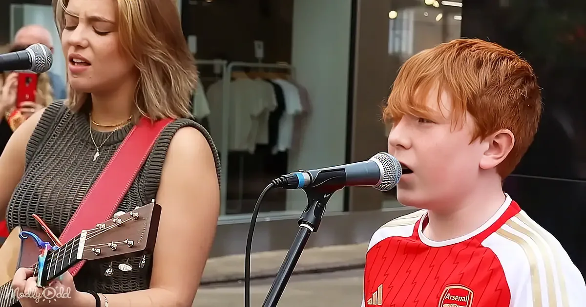 A young boy in a red and white jersey sings into a microphone while a woman plays a guitar nearby.