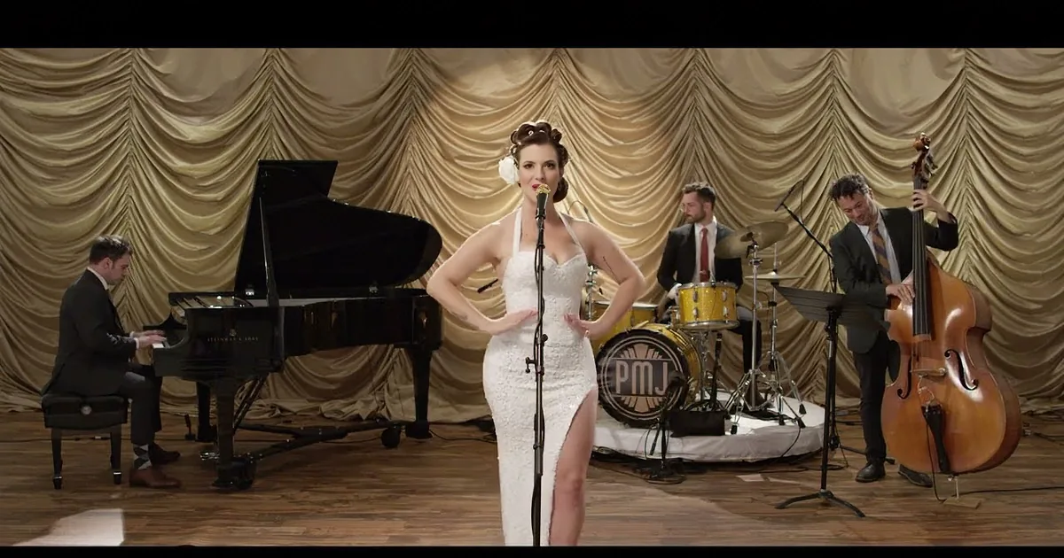 A woman in a white dress sings into a microphone, backed by a jazz band on stage.