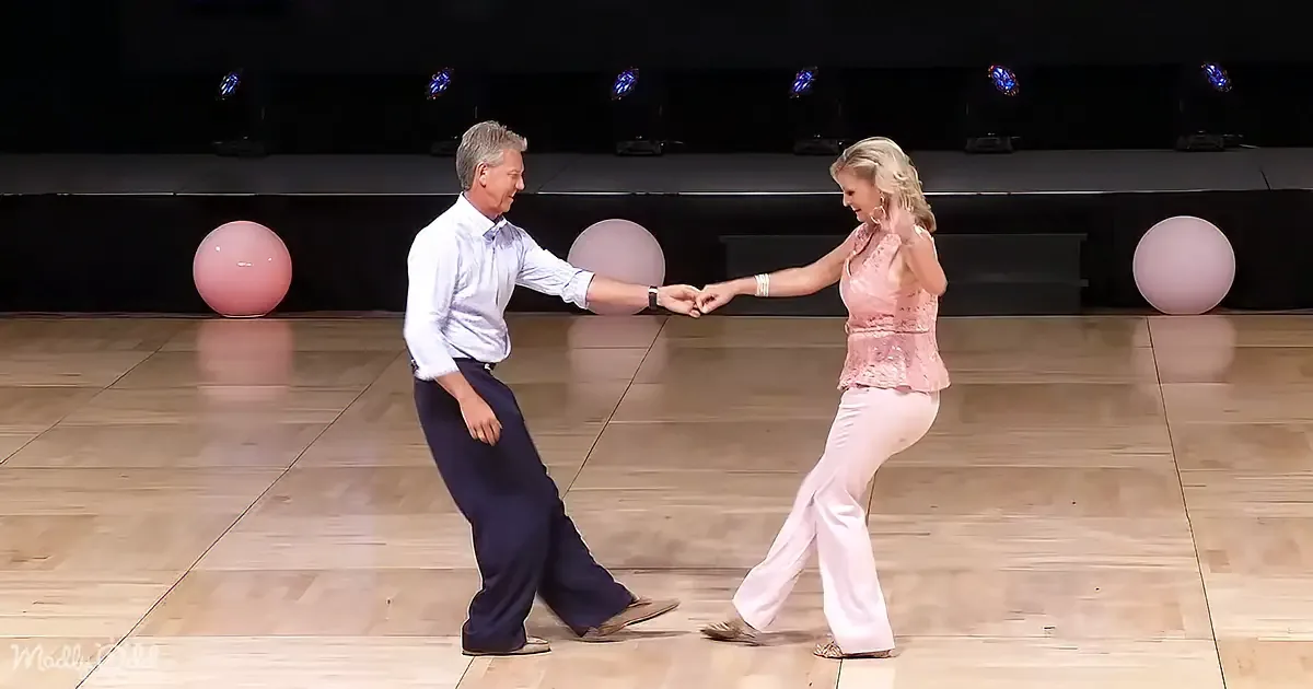 A couple dances gracefully on a wooden floor, holding hands, with a backdrop of pink balloons and stage lights.