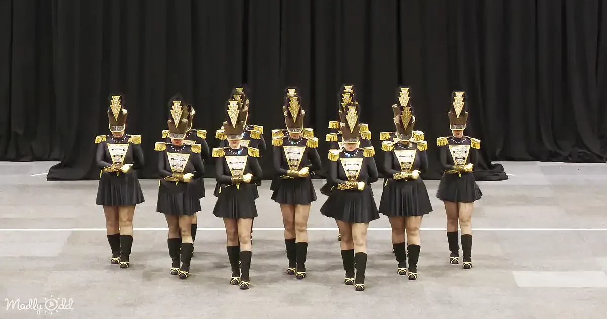 A group of uniformed performers in black and gold costumes stand in a line, holding instruments.