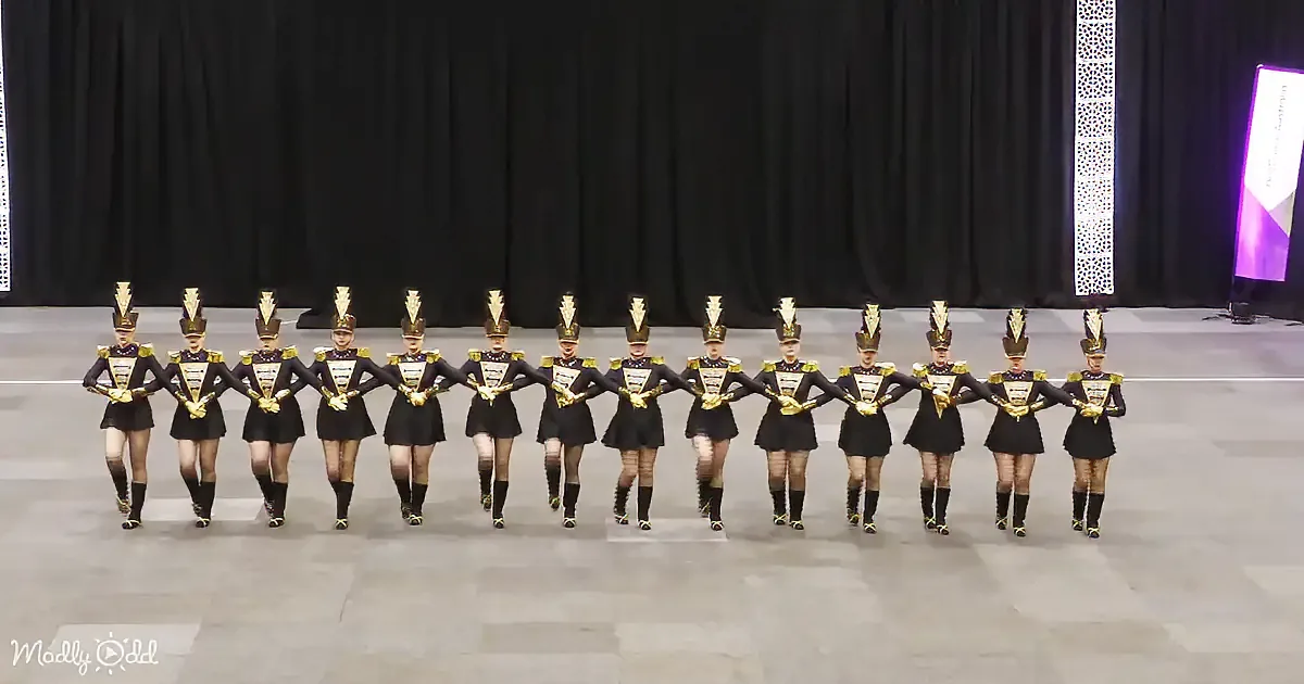 A group of uniformed performers in black and gold costumes march in a line on a stage.