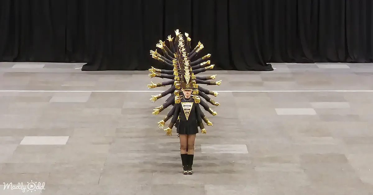 A person in a striking black and gold costume with multiple arm-like extensions stands on a checkered floor.
